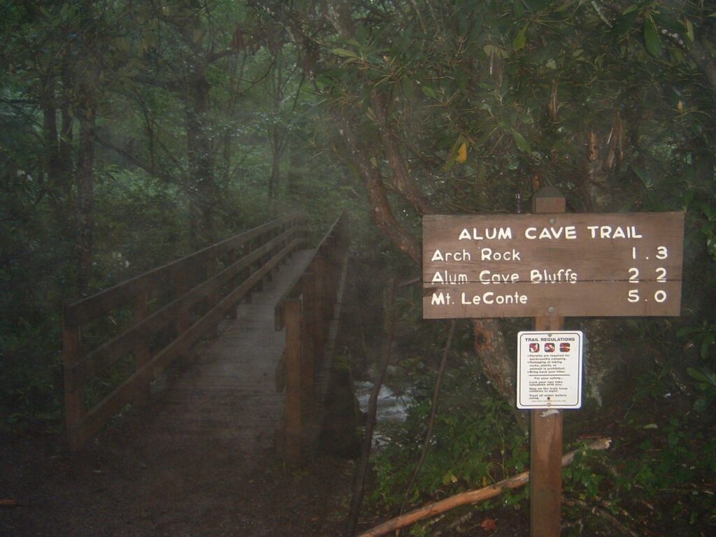 Wooden trailhead sign for the Alum Cave Trail listing mileages to Arch Rock, Alum Cave Bluffs, and Mount LeConte, at a footbridge in fog