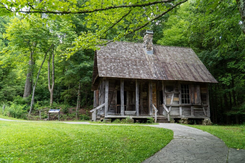 A preserved historic wooden homestead cabin with a stone chimney, set among green trees with a footpath leading to it