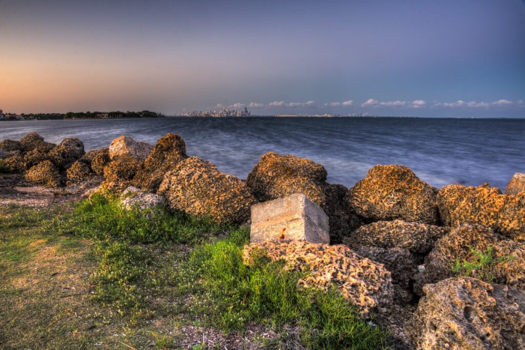 Rocky shoreline along Biscayne Bay with the downtown Miami skyline across the water at sunset
