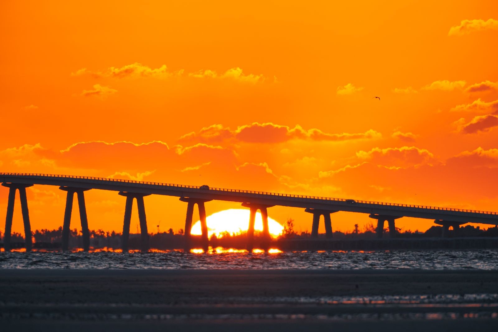 A long causeway bridge stretching across the water at sunset, with an orange sky reflected below.