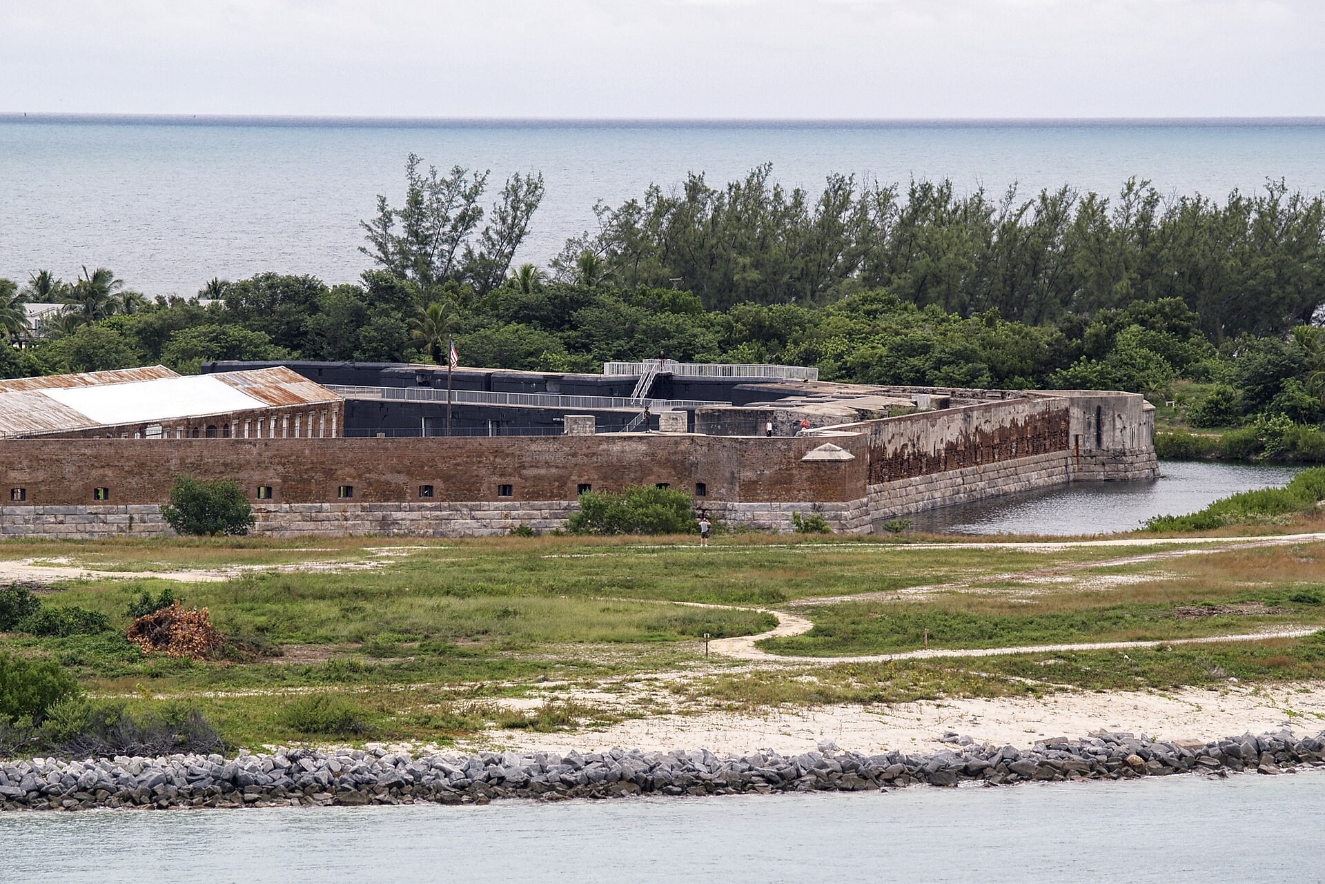 Low brick walls of Fort Zachary Taylor seen from the water, with the rock groin and grass in front and trees behind