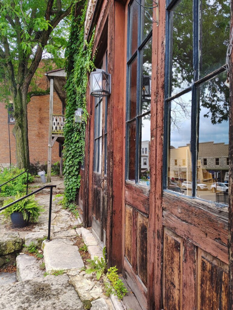A weathered wooden storefront with ivy climbing the wall, brick buildings visible behind it on a hillside street in Galena, Illinois