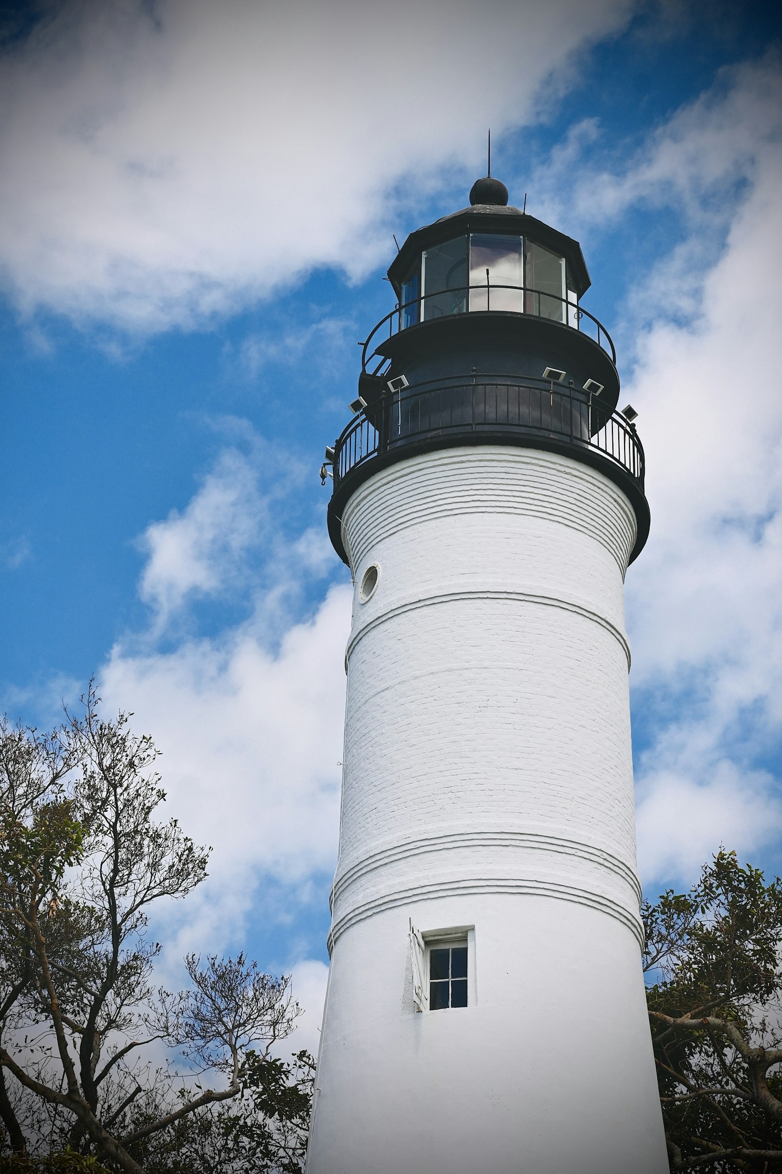 The white brick tower of the Key West Lighthouse with its black lantern room and gallery railing, framed by tropical trees against a blue sky