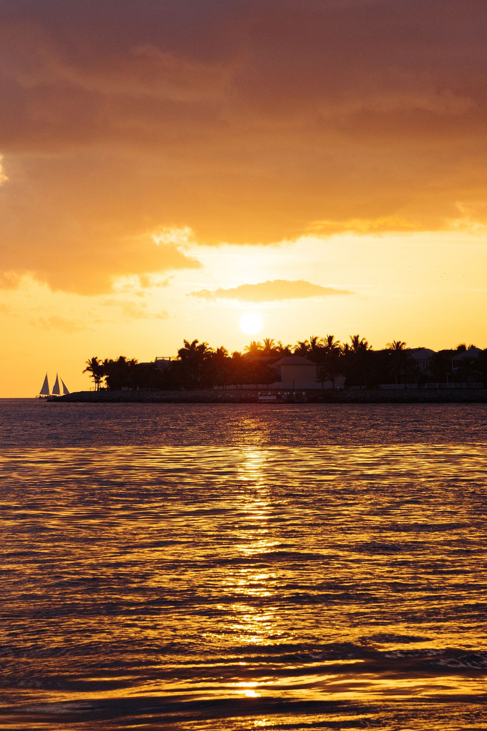 The sun dropping behind a palm-lined Key West shoreline at sunset, with a two-masted sailing boat out on the water and gold light across the harbor