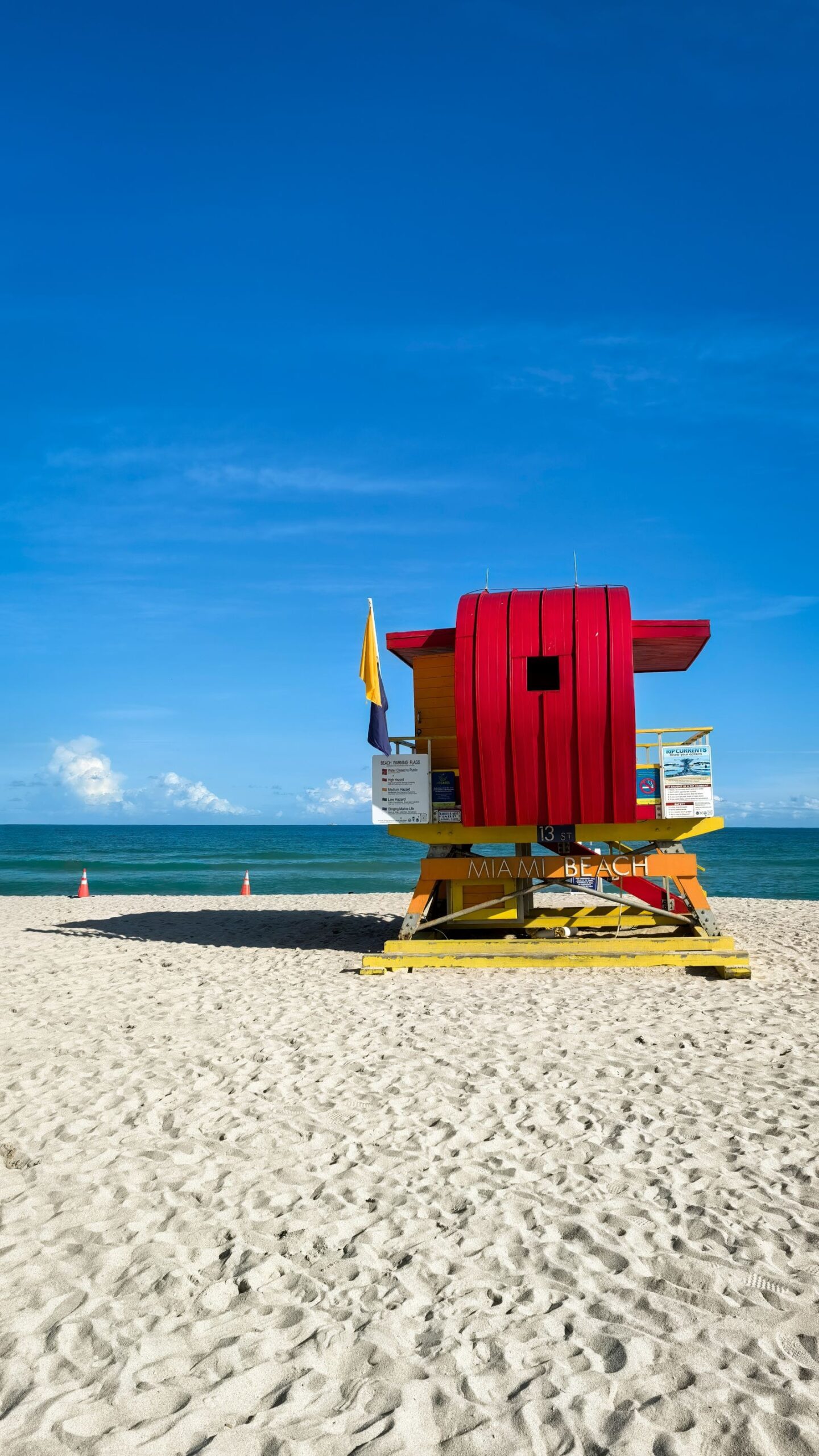 Red and yellow Miami Beach lifeguard tower on the sand, flying a yellow flag above a purple flag