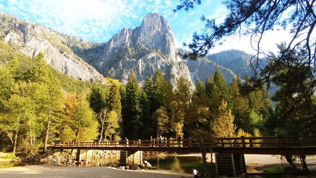 A wooden footbridge over a river in Yosemite Valley with granite peaks and fall-colored trees rising behind it
