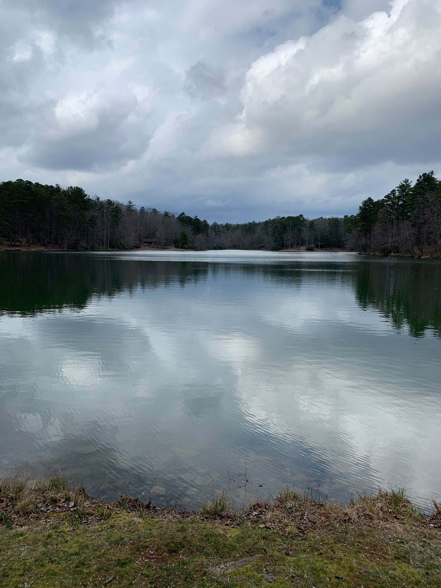 A calm lake at Oconee State Park, South Carolina, under a grey sky, ringed by bare hardwoods and pines, with clouds reflected in the water