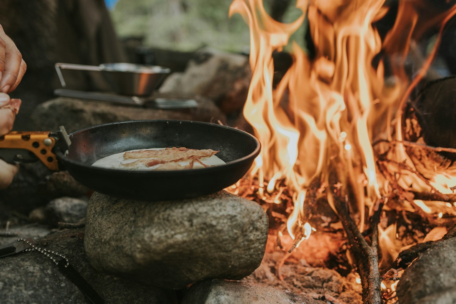 A person cooking food in a skillet over a campfire.
