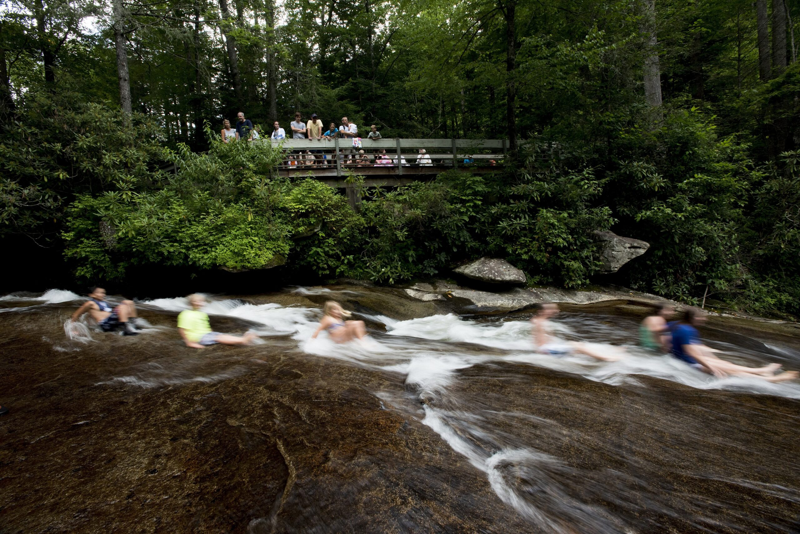 People sliding down the wet granite of Sliding Rock in Pisgah National Forest, with spectators on the viewing platform above