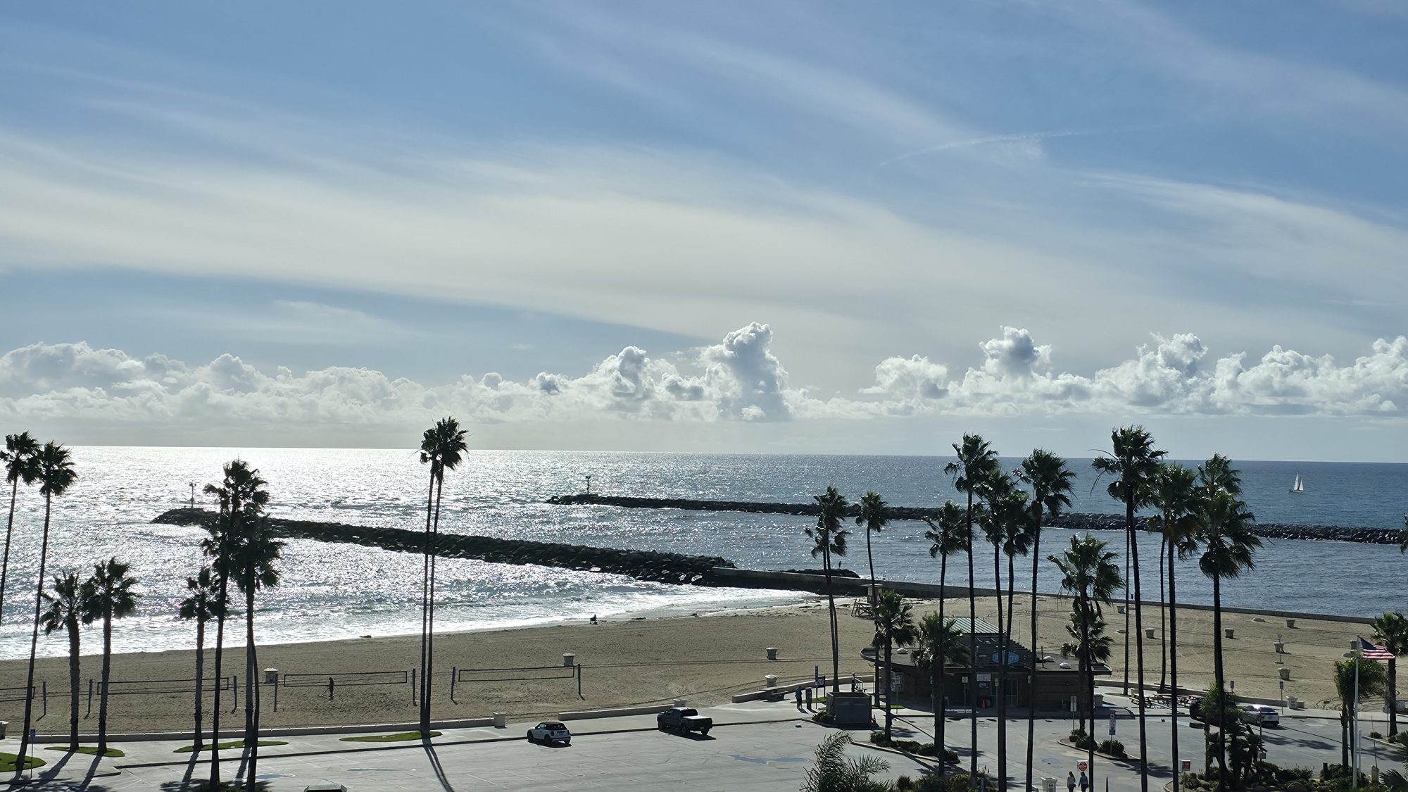 Corona del Mar State Beach, California: the rock jetty, palm trees and a sailboat offshore under wispy clouds