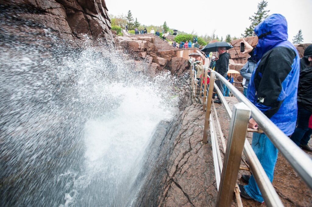 Waves surging into Thunder Hole on the granite shoreline of Acadia National Park as visitors in rain jackets watch from the railing