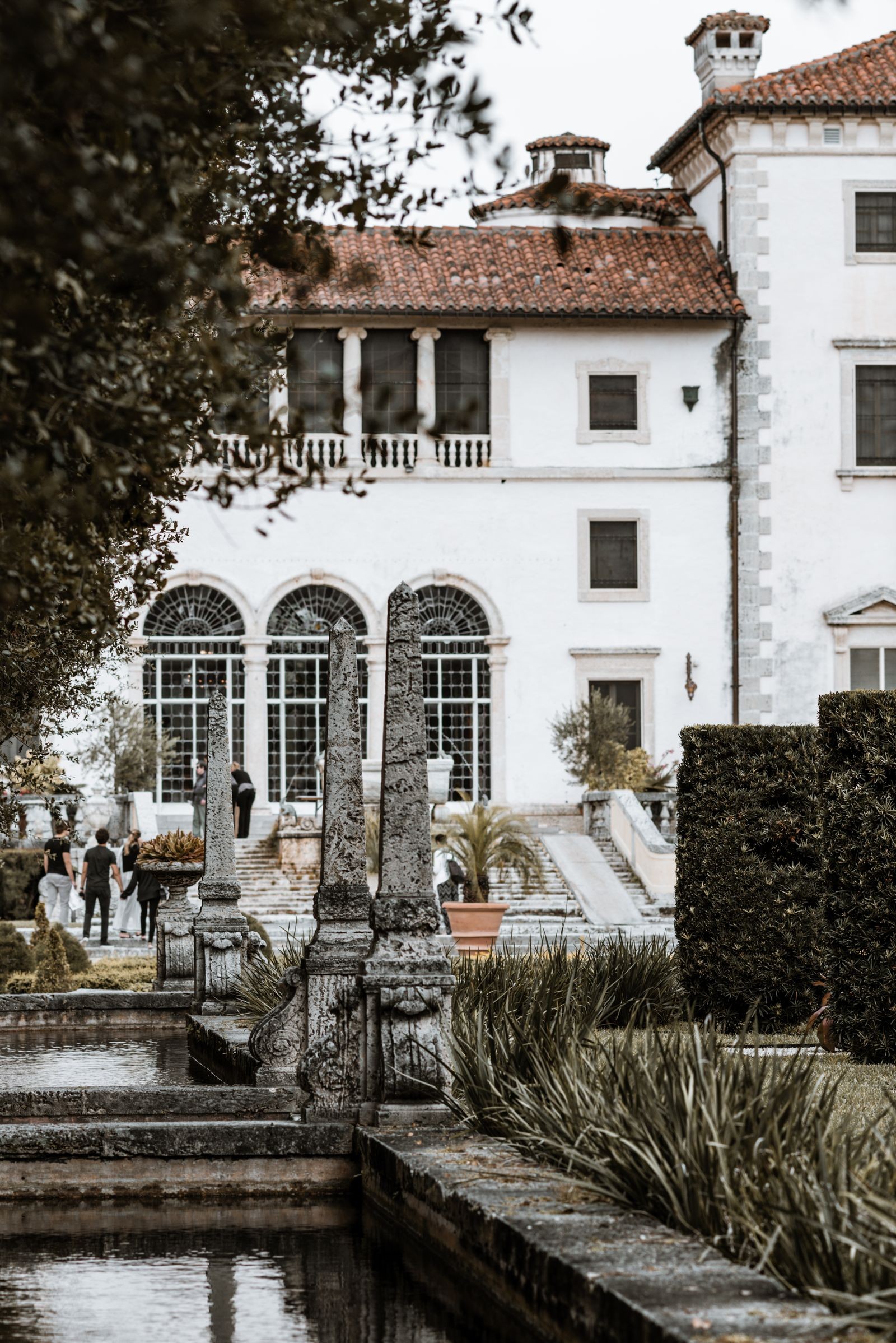 The white stone facade and arched windows of the Vizcaya villa seen across the formal gardens, with stone balustrades and visitors on the terrace