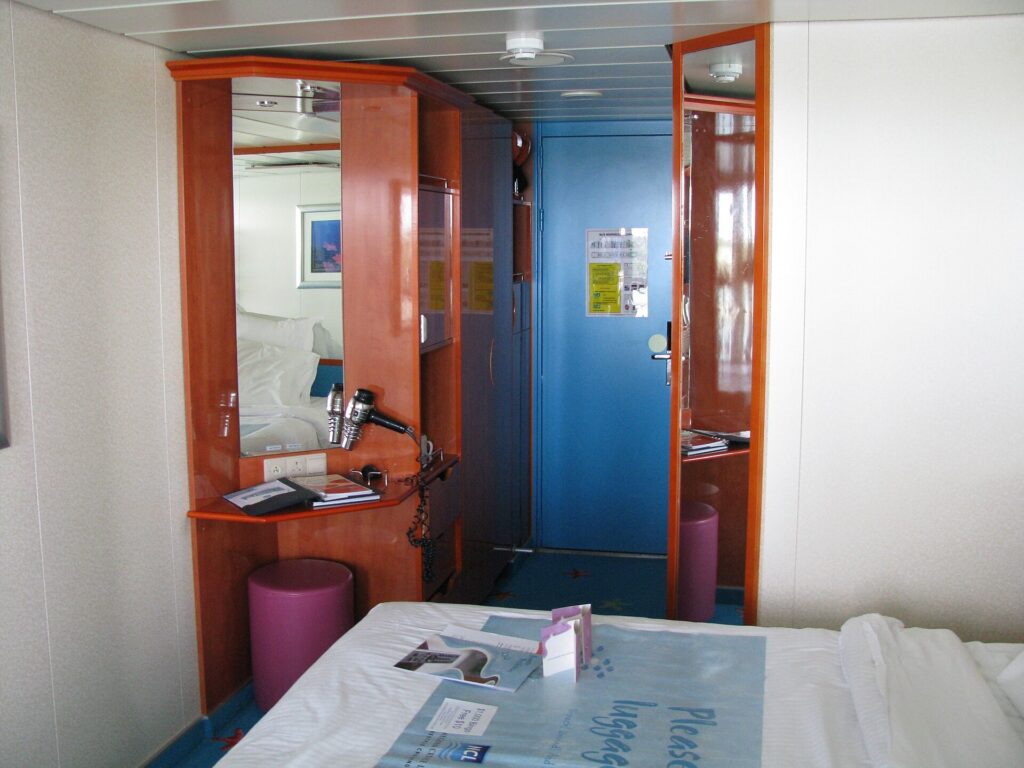 Inside a cruise ship stateroom with a wooden vanity and mirror beside the cabin door and the end of the bed
