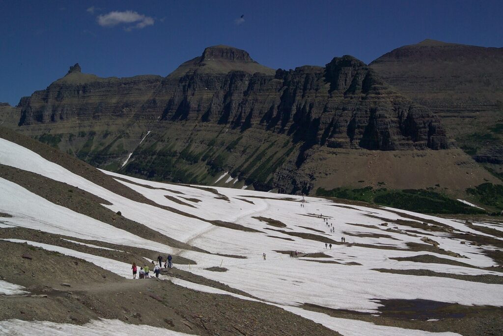 Snowfield below a cliff band with small figures of hikers on the Hidden Lake Overlook trail, Glacier National Park