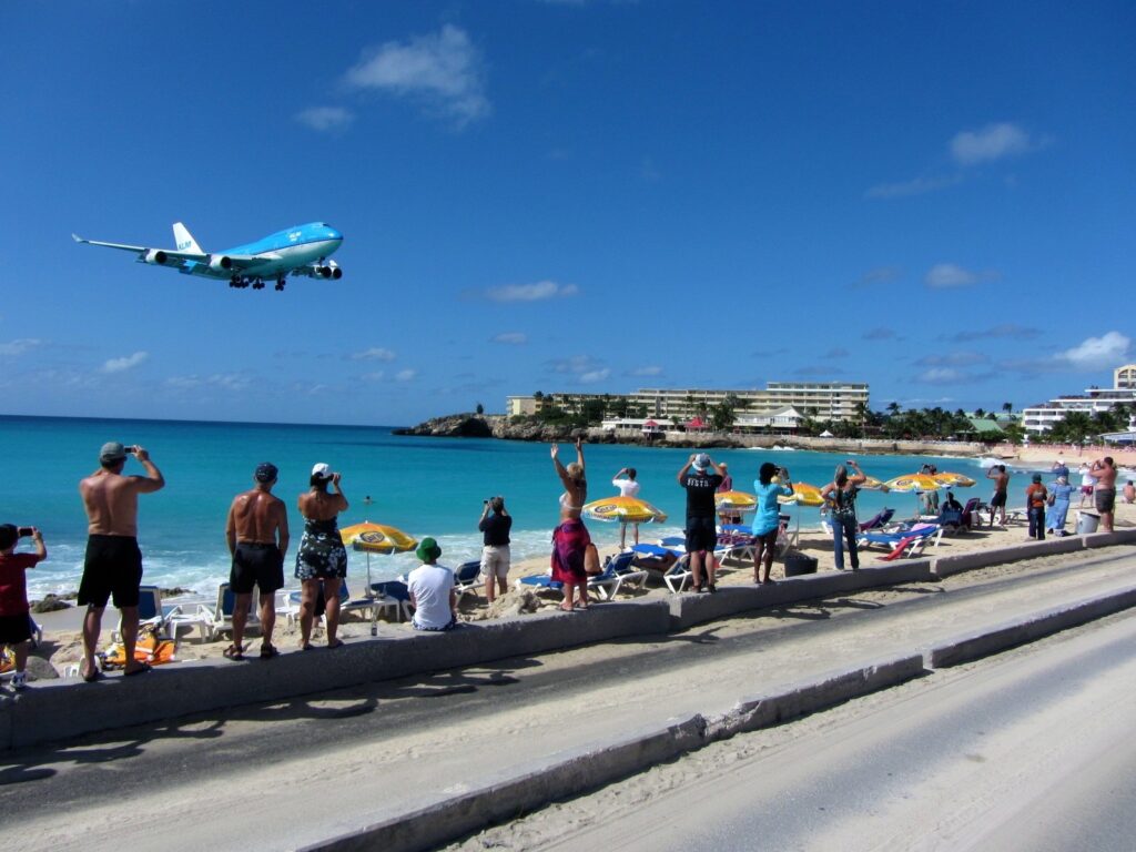 A blue passenger jet on final approach crossing low over the water and spectators at Maho Beach in St. Maarten